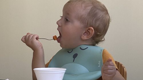 Bebê sentado em mesinha leva garfo à boca na hora da fruta, para da rotina dos bebês no Berçário do Uirapuru