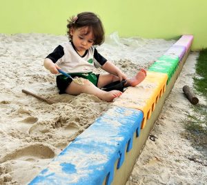 Bebê brinca com colheres na areia do Berçário.