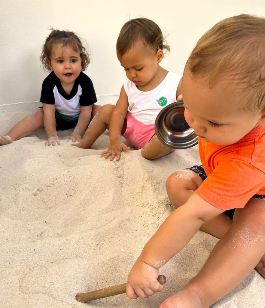 Bebês brincam na areia do Berçário com potes e colheres.