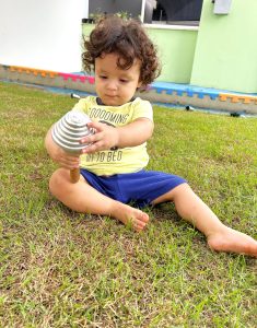 Bebê segura e observa chocalho no gramado no Berçário.