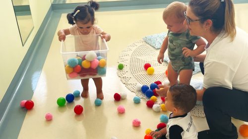 Três bebês que já andam são acompanhados por berçarista em sala de espelhos.