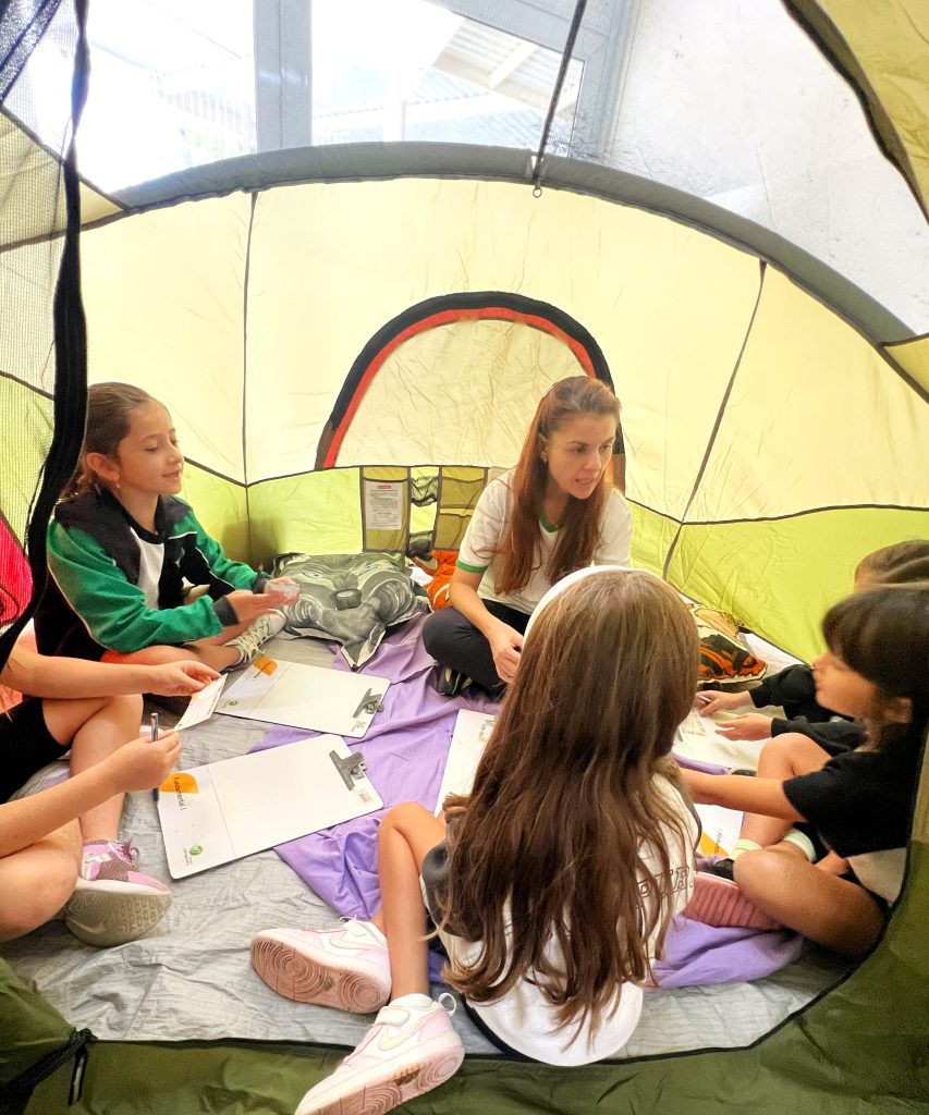 Cinco crianças em roda com professora durante encontro do Clubinho das emoções, do 2º ano, dentro de barraca.